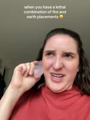 The cover image is a selfie of a fair-skinned woman with dark hair. She looks up and to her right while holding a clear crystal close to her face. Her expression is a mix of amusement and perplexity. The background is blurred, but appears to be a light gray wall. Text overlayed at the top reads, 'when you have a lethal combination of fire and earth placements'.