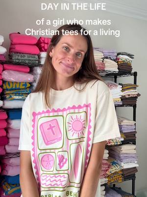 A woman stands in front of shelves filled with folded shirts in a well-lit room. She's wearing a t-shirt with a pink-and-green graphic, smiling directly at the camera. Text overlay says 'DAY IN THE LIFE of a girl who makes Christian tees for a living'.