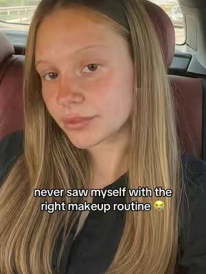 A woman with long blonde hair is seated in a car, facing the camera. The image is well-lit and the background shows the interior of the car.