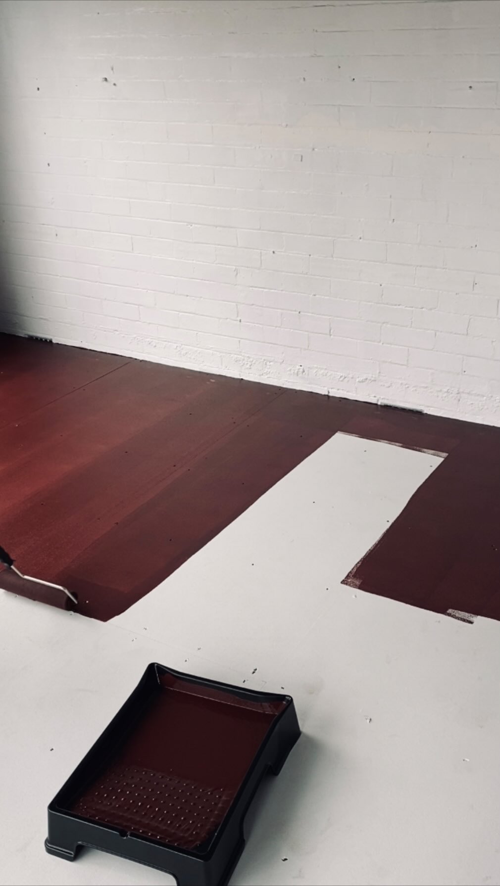 The image is a medium shot showing a room in the process of renovation or painting. The primary colors are red, black, and white. The floor is mostly painted a deep red, with a slightly uneven application, and an opaque white section with an irregular edge. Above, a brick wall is painted white. In the foreground there is a black paint tray filled with the red paint.
