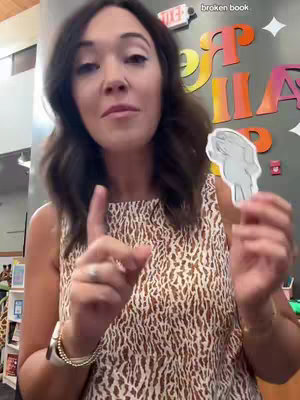 A woman with dark hair is pointing while holding a small cutout in front of a colorful background. She is wearing a patterned sleeveless top and jewelry, standing in what looks like a library or classroom.