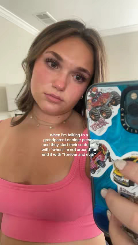 A young woman is taking a selfie, holding her phone in front of her, and looking at the camera with a somewhat sad or thoughtful expression. Text overlay at the bottom provides context. The background is blurred, but appears to be an indoor setting. Her phone case has stickers on it.