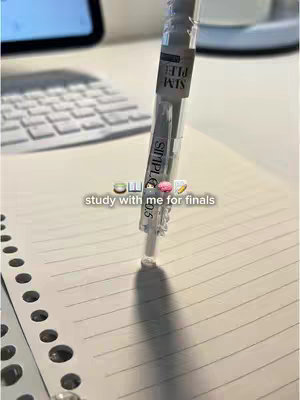 A close-up of a pen, positioned vertically above a lined notebook. The pen is white and clear, with visible ink. Below the pen is the hook text 'study with me for finals'. There is a partially visible white keyboard and a corner of a black electronic device in the background. The composition is clean, with a focus on the pen and the text.