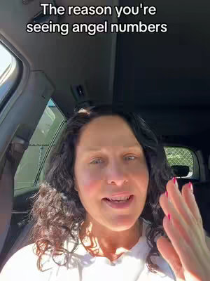 A woman with long, dark, curly hair is seated in a car, looking directly at the camera and speaking. The image is a selfie taken from a slightly low angle. She has fair skin, and her expression is open, with a slight smile. The background of the car suggests an outdoor setting, with sunlight filtering in. Text overlayed at the top reads, 'The reason you're seeing angel numbers.'