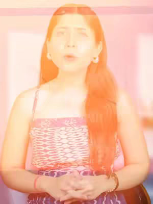 The image shows a woman with long reddish-brown hair. She is wearing a patterned spaghetti strap top and has hoop earrings. Her hands are clasped in front of her. There is a bright orange overlay that makes the image a little blurry.
