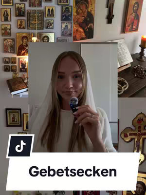 A woman dressed in white holds a microphone, standing in front of a wall decorated with religious icons and a crucifix. The bottom of the image has a white banner with black text.