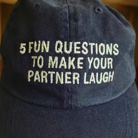 A close-up shot of a navy blue baseball cap. The hat is angled, and the white embroidered text, "5 FUN QUESTIONS TO MAKE YOUR PARTNER LAUGH," is clearly visible. The background is out of focus, with hints of a wooden surface.