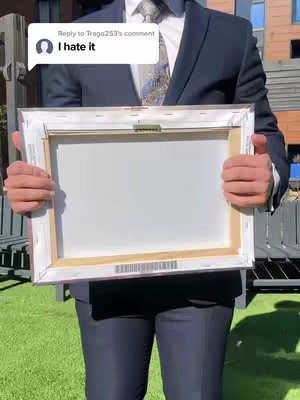 A person in a dark suit holding a white canvas with a wooden frame. There is a comment bubble overlay with the text "I hate it". The person is standing outside, and wearing a tie.