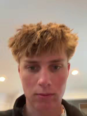 A close-up shot of a young man's face, seen from the chest up. He has short, light brown hair and a neutral expression. The background is blurred, suggesting an indoor setting with overhead lighting and a white ceiling. The man is wearing a dark-colored jacket or shirt.