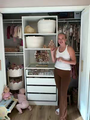 A woman stands in front of a well-organized white closet that features open shelving, drawers, and clothing racks. In the background, there's another closet, filled with hanging clothes. A baby plays and a toy piano are next to the main closet. The woman points to items.