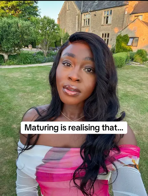 A woman with long, wavy black hair is centered in the frame, looking towards the camera. She has a soft, warm skin tone, with a hint of lip gloss. She is set against a lush green background with a large stone building. There's text overlaid at the bottom of the image.