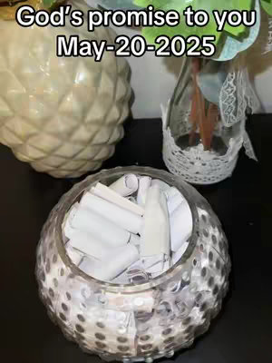 The image shows a glass container filled with rolled-up white papers. The container is placed on a surface in front of other blurred objects.