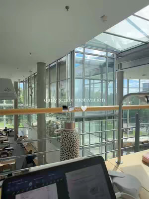 An interior shot of a modern study space, likely a library or university building. Natural light streams in through large glass windows and a glass ceiling, illuminating study desks and a walkway. In the foreground is a laptop with an open screen, and a water bottle in a patterned sleeve. The text overlay reads 'daily study motivation'.