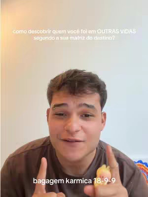 A young man with short brown hair looks directly at the camera with a slightly open mouth, raising his index fingers to point upward. Light beige text, asking how to discover past lives, is written above his head. Brown text stating 'bagagem karmica 18-9-9' is placed near his chest. He is wearing a brown t-shirt with some colorful decor in the background. His eyebrows are pierced, there is a ring on his finger and a yellow decoration on his other index finger.