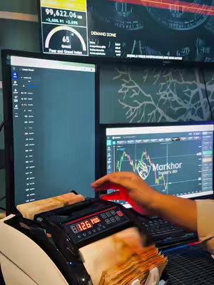 The image shows a desk setup with multiple computer screens displaying financial data, with a hand counting money in the foreground. A cash-counting machine is also visible.