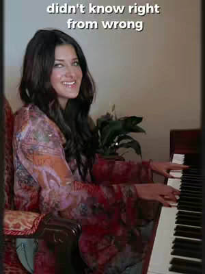 A woman with long dark hair is seated at a piano, smiling. She is wearing a floral top. Text appears above her head.