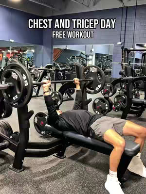 A man is lying on a weight bench in a gym, lifting a barbell overhead. Text overlays at the top read 'CHEST AND TRICEP DAY' and 'FREE WORKOUT'. The gym has mirrors and weight-training equipment visible in the background. The man is wearing gym attire, gray shorts and black shirt and sneakers.