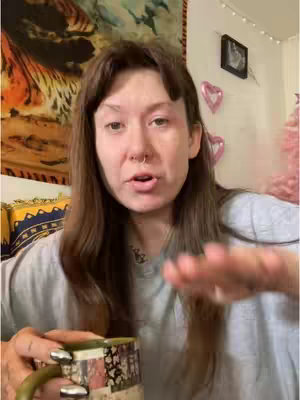 A woman with brown hair and bangs looks directly at the camera. She holds a colorful mug and gestures with her right hand. Her face has minimal makeup, with a nose ring. A backdrop of various items, including a tapestry, a framed picture, and string lights, adds to the setting.