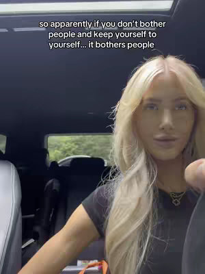 A woman with long blonde hair sits inside a car, looking towards the camera. Text overlays the top of the frame.