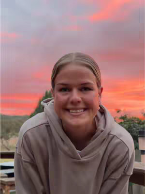 A young woman with blonde hair and fair skin smiles at the camera in the foreground. She wears a tan hoodie. The background is a sunset with a gradient of pink and orange.