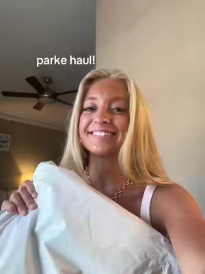 A blonde woman is smiling and holding up a white package. She is wearing a necklace and a light-colored top, with a ceiling fan in the background.