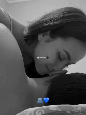 A black and white photo shows two people intimately close. One person, partially visible, is leaning over the other and kissing them on the forehead. The person in the foreground is slightly blurred, while the person on top is clearer. The lighting is soft, and the background is indistinct.