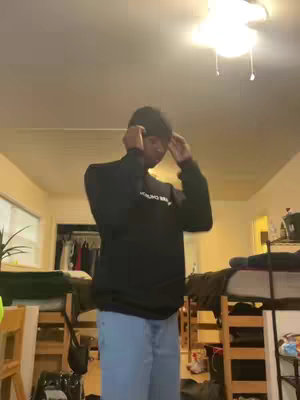 A person is standing in a room, wearing a black sweatshirt and jeans, appearing to adjust their hat. The room is a basic dorm or apartment setting with a bed, closet, and a light fixture on the ceiling.