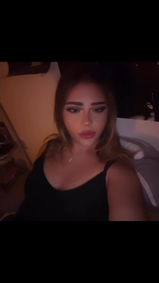 A close-up shot of a person with heavy makeup. The person is wearing a black tank top and has long, light brown hair. The background is blurred and dark.