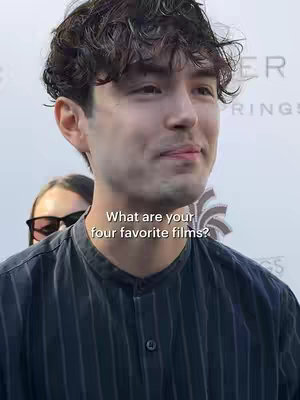 A medium close-up shot features a man with dark, curly hair, turned slightly to the side, looking towards the right. The man is wearing a dark, striped shirt. Overlayed on the image is the text asking, "What are your four favorite films?" Behind him, another person's face is partly visible. The background appears to be a light, outdoor setting.