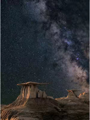 The cover image is a vertical shot of a night landscape, featuring towering rock formations under a starry sky. The foreground is dominated by the detailed, textured rock structures, with a warm, earthy color palette of browns and tans. The background showcases a deep blue sky filled with countless stars and the blurred glow of the Milky Way, creating a contrast with the darker rocks. The overall composition emphasizes the vastness of space and the intriguing shapes of the rock formations.