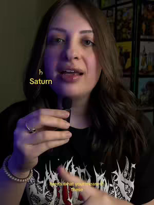 A woman with long brown hair is centered in the frame, wearing a black t-shirt. The image has text overlays in yellow, including an astrological symbol and the word "Saturn." The background is softly lit, with a hint of other objects or visuals.