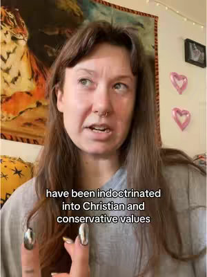 A woman with brown hair is centered in the frame, speaking to the camera. Text overlayed at the bottom of the screen reads 'have been indoctrinated into Christian and conservative values'. The background includes a colorful tapestry, a wall-mounted photo, and two heart-shaped balloons.