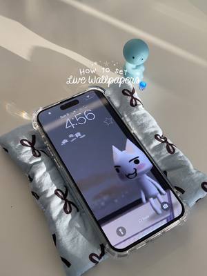 A picture on a white surface featuring a phone displaying a cartoon character cat wallpaper laying on a blue cushion. There is a small blue figurine standing upright behind the phone and a text overlay reading: "How to set Live Wallpapers".