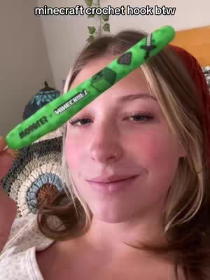 A young woman with fair skin and blonde hair is holding a vibrant green crochet hook with a Minecraft design, showcasing it towards the camera. The hook is the primary focus, with the woman's face partially visible, showing a relaxed and friendly expression. The background contains subtle details.