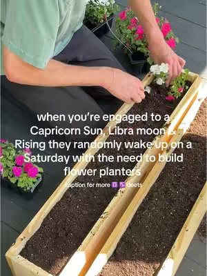 A person is shown from chest up, kneeling down while planting flowers in a long wooden planter box. The background includes a second planter box, flowers in pots and a wooden deck.
