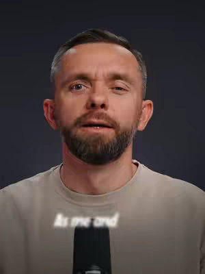 A medium shot of a man with short, dark hair and a beard, centered in front of a dark background. The man is looking towards the upper left with a slight frown, and the text overlay is semi-transparent and white.