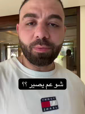 A man with a beard and a white t-shirt is centered in the frame, looking towards the camera. Below him is a black box containing Arabic text. Behind him is a blurred background of a room with a reflective door.