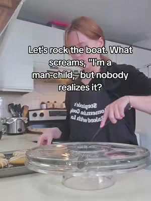 The image shows a woman in a kitchen setting. She stands behind a countertop. There's a glass cake stand in front. She appears to be holding a spatula. Background includes a stove and cabinets.