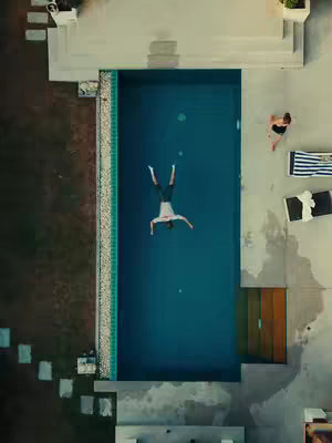 An aerial shot shows a man in a body of water, with a person lounging by the side of the pool. The pool is rectangular, with the man in the center. A person is positioned on the upper right corner of the image on a lounge chair. The background is a concrete surface and grass.