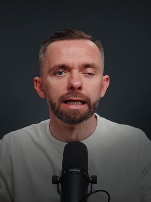 A medium shot of a man with short brown hair and a neatly trimmed beard, looking directly at the camera with a serious expression. He is wearing a white t-shirt, and a black microphone is positioned in front of him. The background is a plain, dark gray color, focusing attention on the speaker. The lighting is moderate, highlighting the details of his face and the microphone.