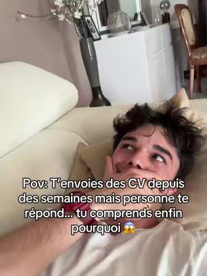A selfie-style shot of a young man lying on a sofa, looking upwards with a stressed expression, hand covering his mouth. The background has a neutral tone with interior elements softly visible.