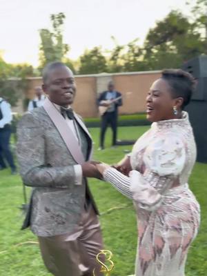 A couple in formalwear dancing and holding hands outdoors on a sunny day. The man is wearing a gray suit and the woman a patterned dress. A guitarist plays in the background. The couple faces the viewer in the center of the frame with an unfocused background of greenery.