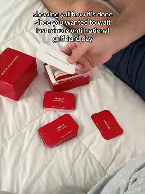The cover image is a top-down shot of a bed with a white comforter. Scattered across the bed are several red boxes and cards from a game. One hand, presumably the creator's, is holding a white card from the game, partially obscuring the view of the items on the bed. The image is well-lit, with natural light illuminating the scene. The overall composition suggests an informal, relaxed setting.