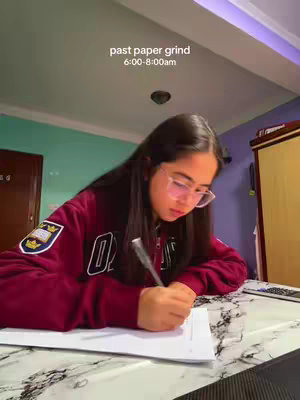 A young woman with long dark hair and glasses is seated at a table, writing on a paper. The room has teal and purple walls, and the lighting appears to be a soft, natural light. She is wearing a maroon hoodie with a logo on the left breast. Text overlay in the upper center says "past paper grind 6:00-8:00am".