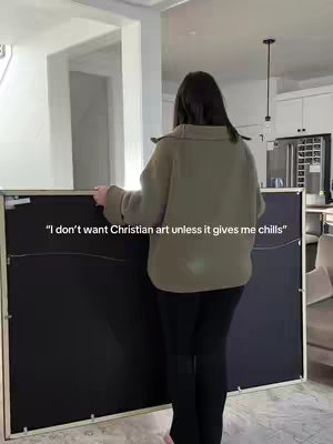 A woman stands facing away from the camera, holding a large framed art piece, likely a canvas or print, in front of her. She is wearing a light green colored sweater and black leggings. The background shows a well-lit interior space with white walls and a glimpse of a kitchen.