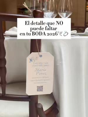 The image shows a close-up of a wedding reception table setting. A wooden chair has a tag hanging on the back, with text in Spanish, a QR code, and a small floral emblem. The table is covered in a white tablecloth, with the bottom part in the background blurred.