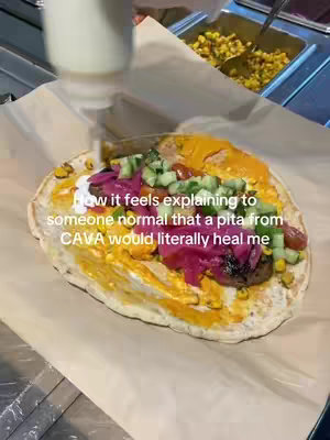 A close-up shot of a pita sandwich, filled with various ingredients such as colorful toppings, sauces, and what appears to be meat or falafel. The sandwich sits on a white parchment paper, with more food items visible in the background.