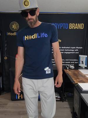 A man wearing a cap, sunglasses, and a navy blue t-shirt that says 'HodlLife' stands in front of a black background with white text. The man is looking down with a serious expression. He is wearing sunglasses, and has a beard. His t-shirt is tucked into light-colored pants. There are logos and additional text in the background. The pose is static and the overall composition is casual.