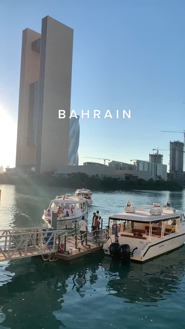 A scenic view of a harbor in Bahrain, featuring several white boats docked at a pier. A modern skyscraper and other buildings comprise the backdrop, with a clear blue sky overhead. The composition highlights the city's skyline and waterfront.