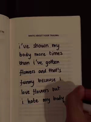 An open book with handwritten text on a page titled 'WRITE ABOUT YOUR TRAUMA'. The handwriting is in black ink with uneven lines. A hand, likely the author's, is visible at the bottom right of the page. The overall composition is simple, with warm tones and a focus on the handwritten message.